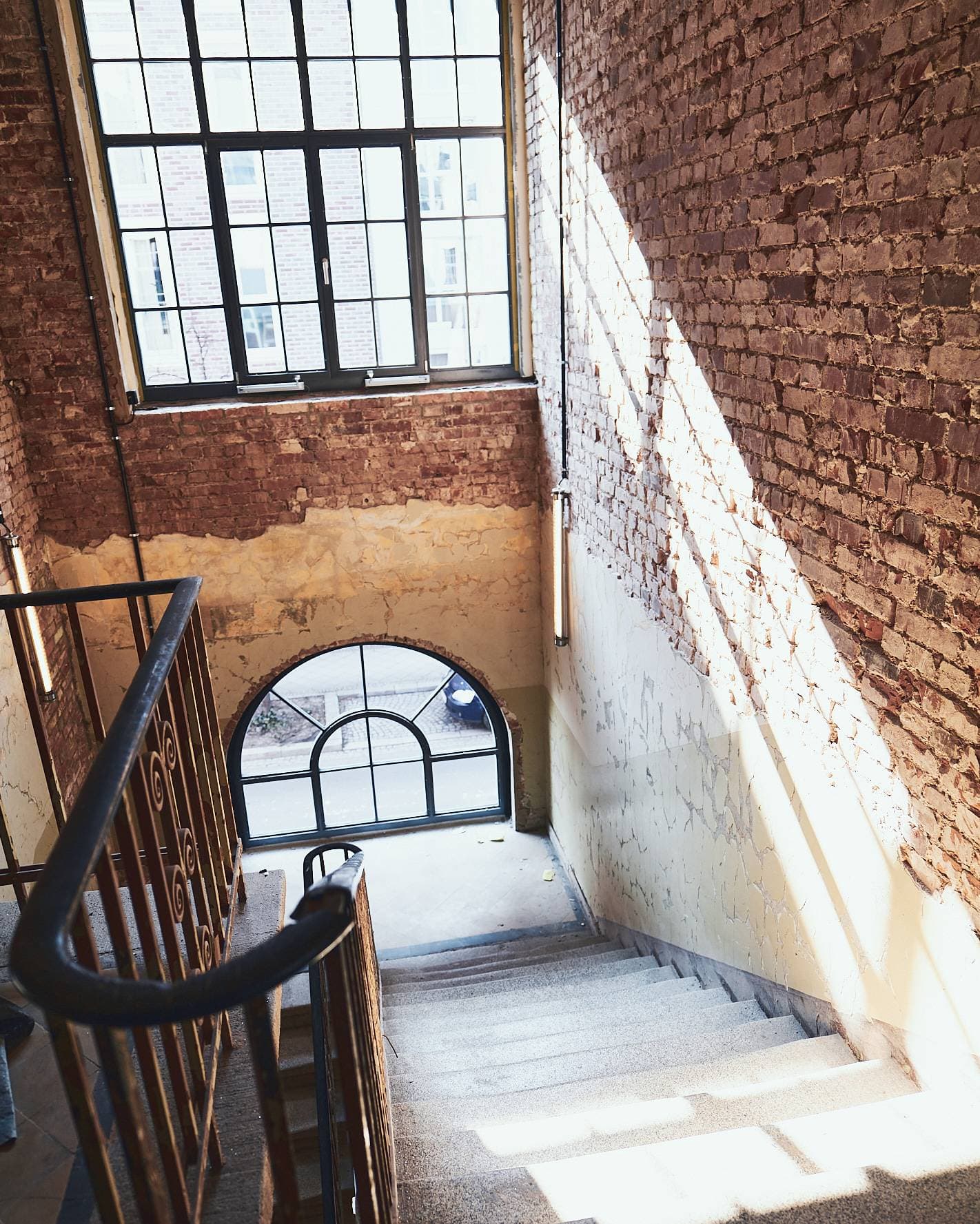 Ein Treppenhaus mit Geländer, Backsteinwand und Rundbogenfenster im Sonnenlicht